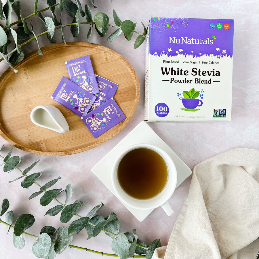 White Stevia Powder Blend Packets box, purple packets on a wooden tray and a cup of tea, with eucalyptus stems around