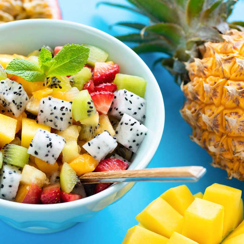 Fresh fruit salad with passion fruit, kiwi, mango and pineapple in a bowl surrounded by pineapple and mango