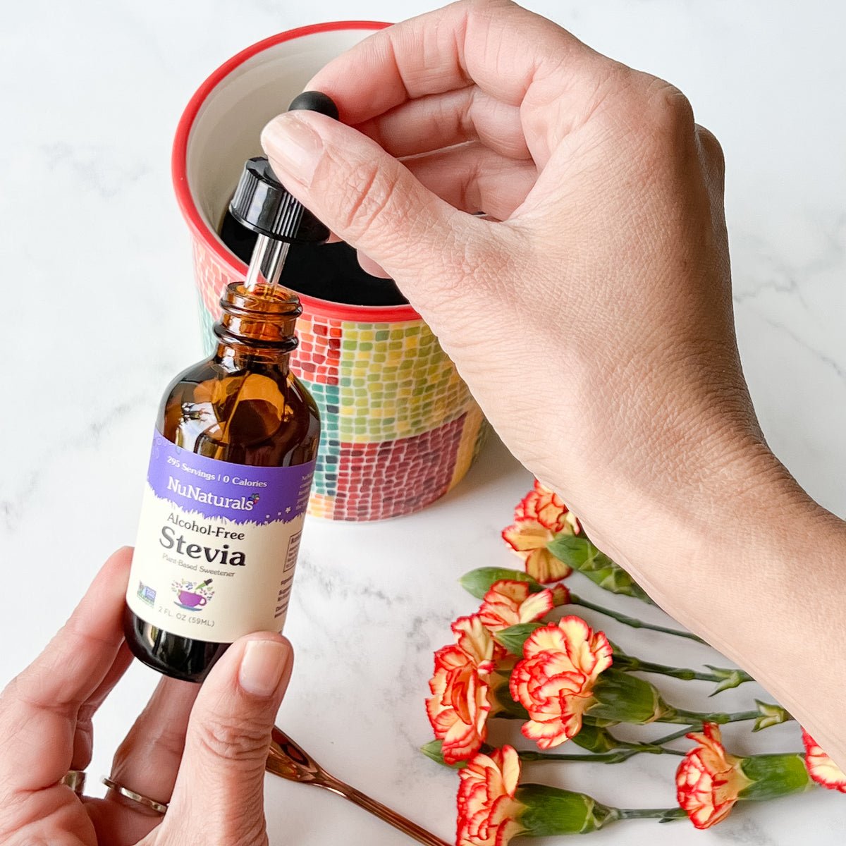 Alcohol-Free Stevia 2 oz dropper bottle held over a mug by one hand, with orange carnations on a marble surface