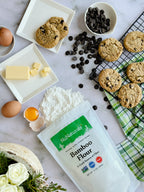 Bamboo Flour bag beside chocolate chip cookies, butter, eggs, a cracked egg yolk and a pile of flour