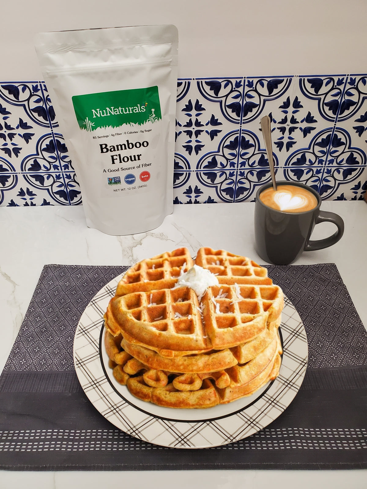 Bamboo Flour bag behind a plate of waffles with cream, next to a mug of latte on a patterned tile counter