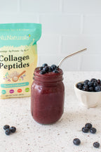 Collagen pouch behind a jar of blueberry smoothie with a metal straw, with a bowl of blueberries beside it