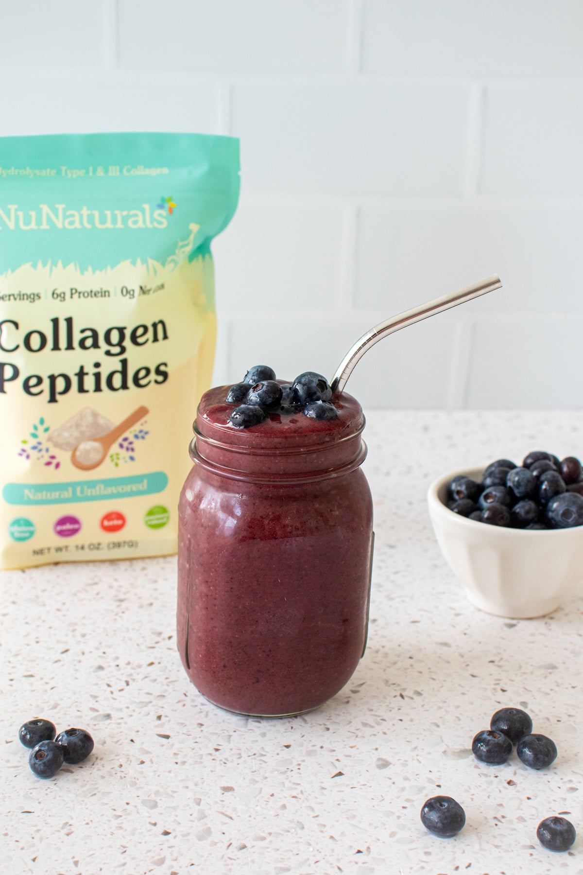 Collagen pouch behind a jar of blueberry smoothie with a metal straw, with a bowl of blueberries beside it