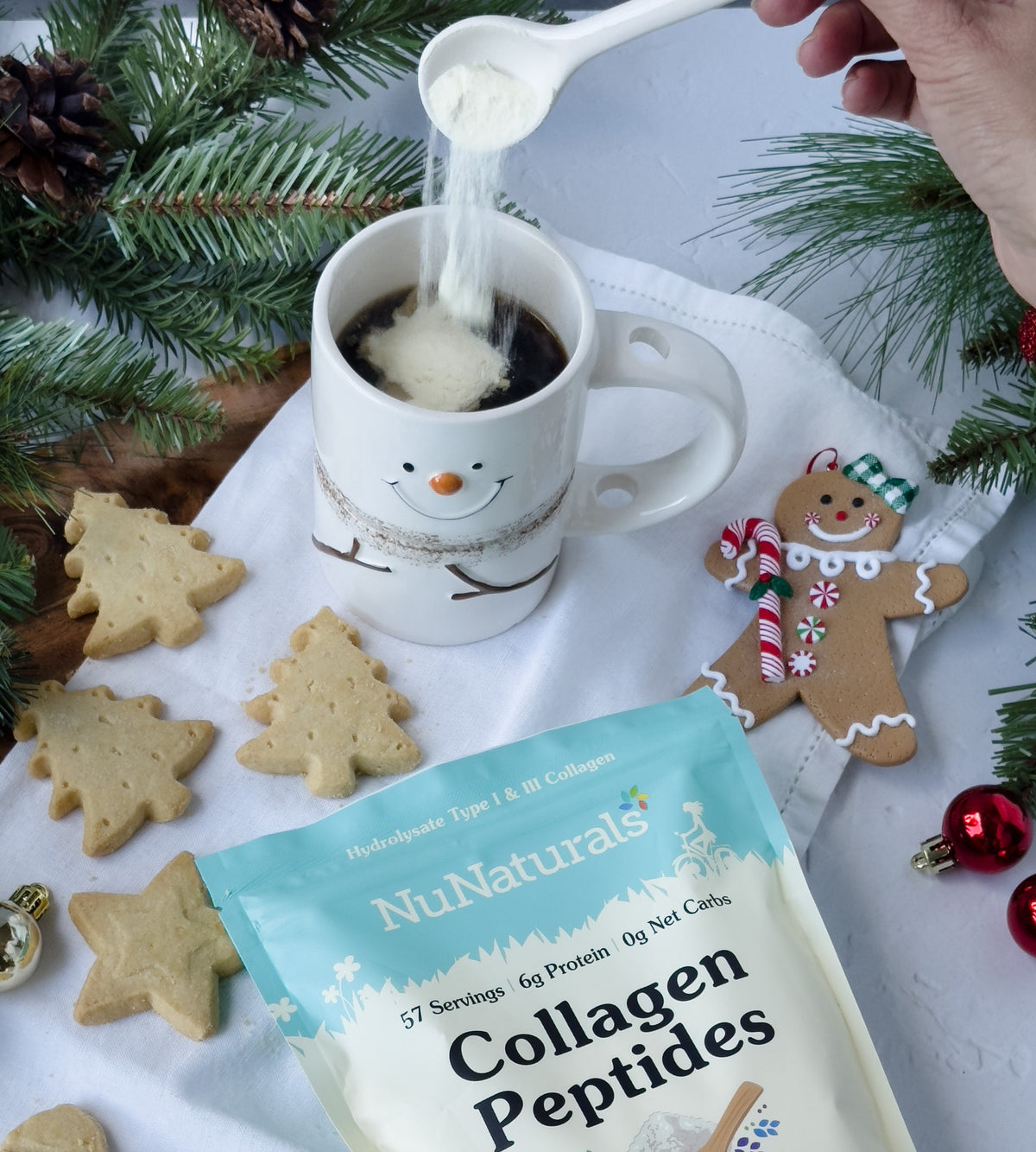 Collagen Peptides pouch at the bottom of the frame as powder is poured into a snowman mug, with cookies and pine branches