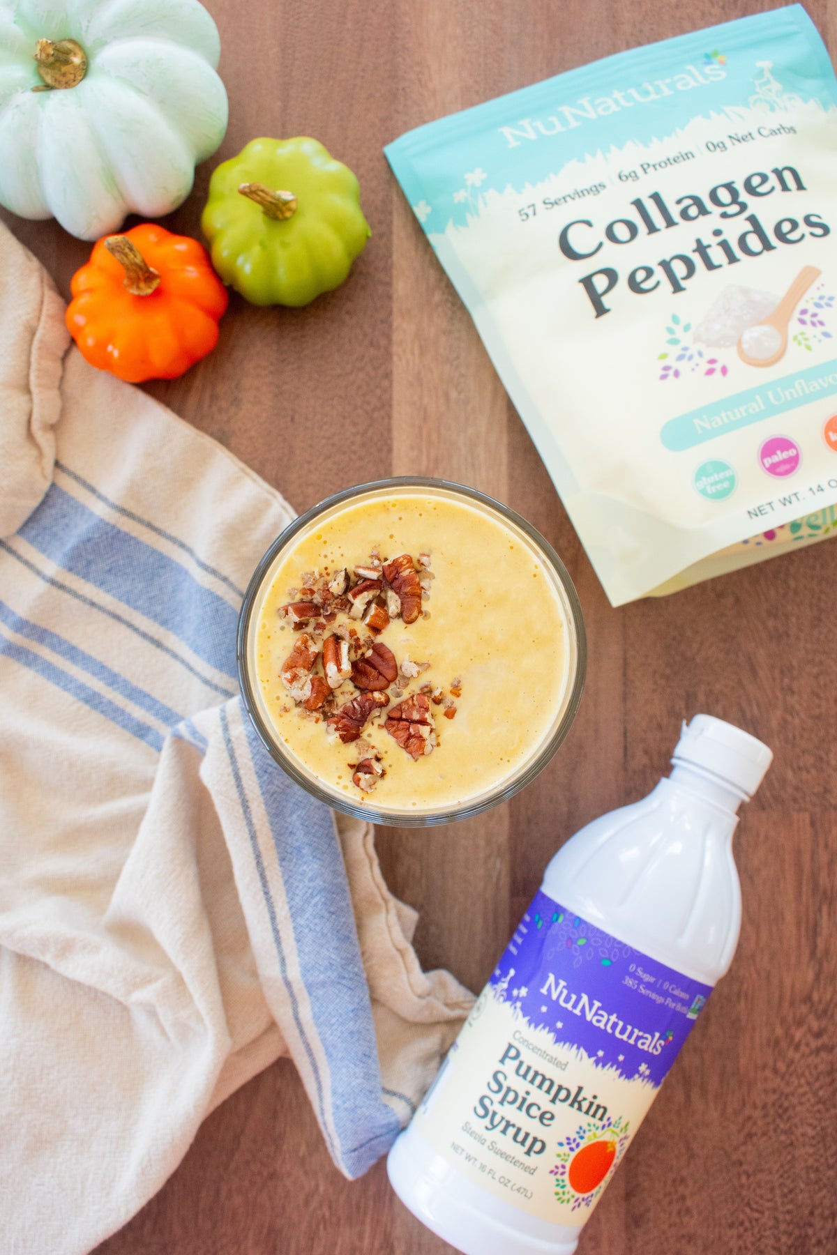 Collagen pouch and a syrup bottle around a glass of golden smoothie topped with chopped pecans, with small pumpkins