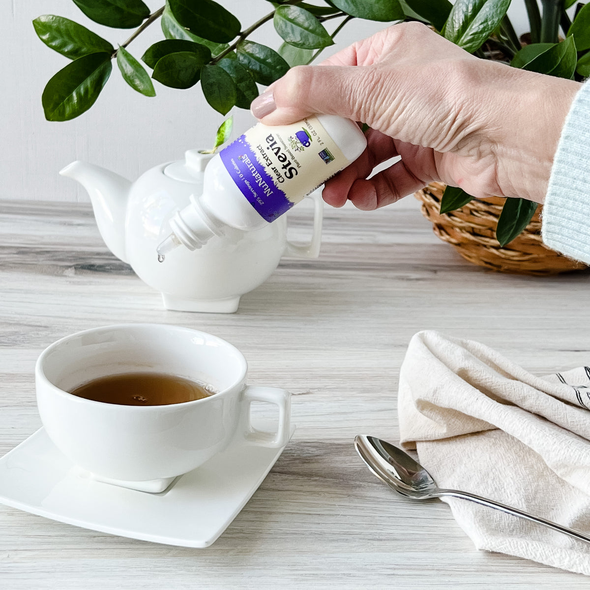Clear Extract Stevia 2 oz dropper bottle held over a cup of tea, with a teapot and spoon on a wood table