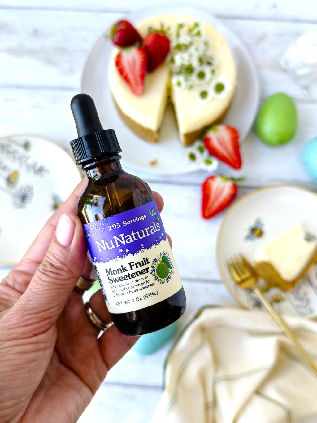 Monk Fruit Sweetener 2 oz dropper bottle held in a hand above a cheesecake with strawberries and Easter eggs
