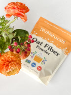 Oat Fiber pouch with front label, lying beside orange flowers and small red berries on a white surface