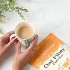 Oat Fiber powder pouch beside a hand-held striped mug of latte, with small yellow flowers on a marble surface