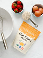 Oat Fiber Powder pouch beside a whisk in a mixing bowl, a measuring scoop of flour, strawberries, and eggs