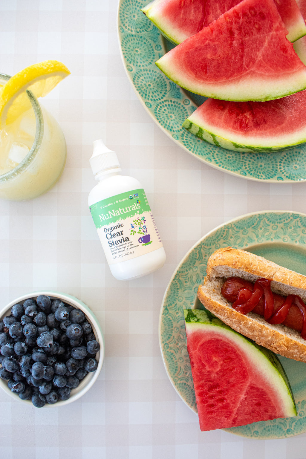 Organic Clear Extract Stevia 4 oz dropper bottle lying on a tiled surface with blueberries, watermelon slices and a hot dog