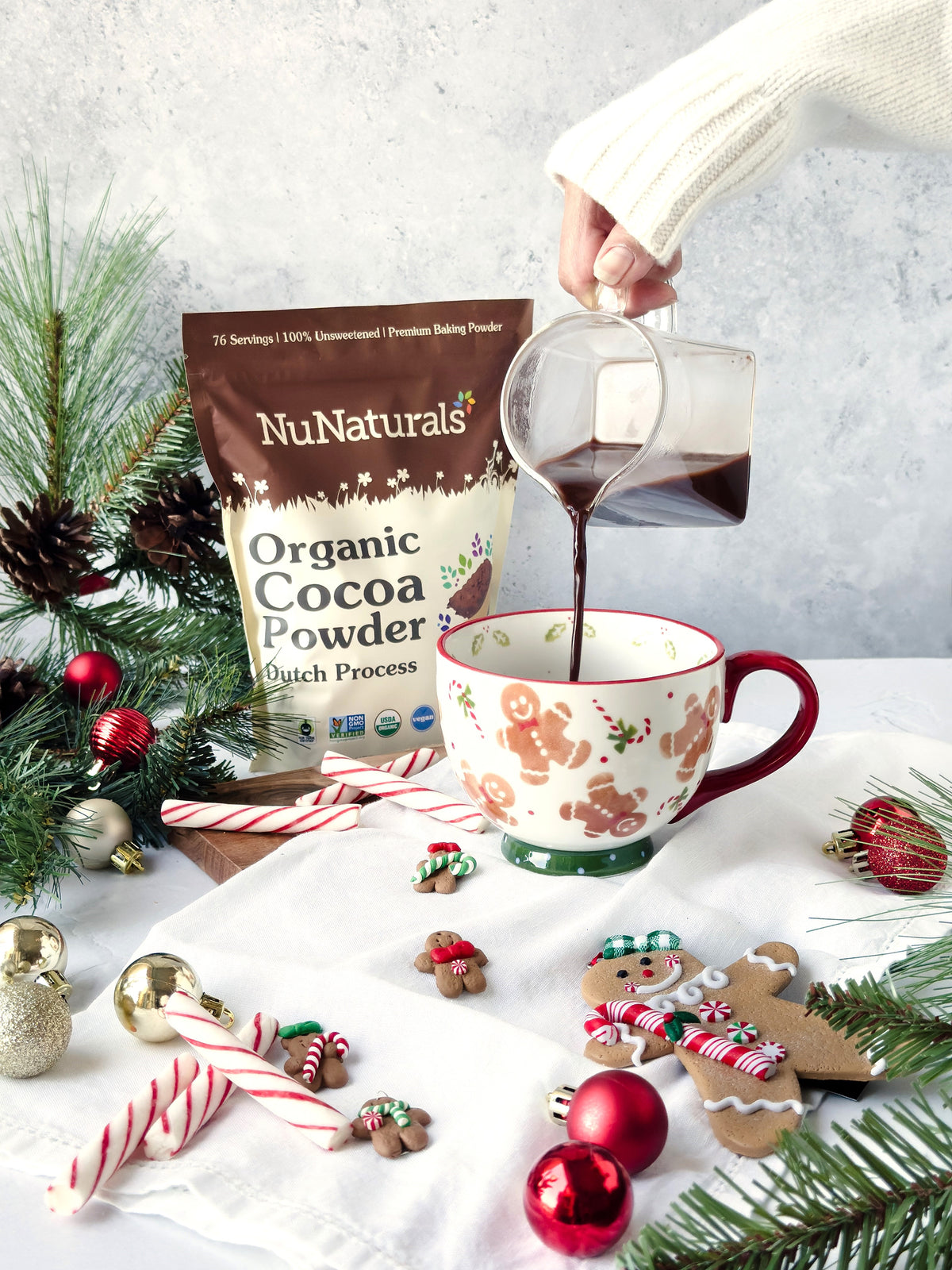 Organic Dutch Cocoa pouch behind a hand pouring chocolate into a holiday mug, with pine branches and candy canes
