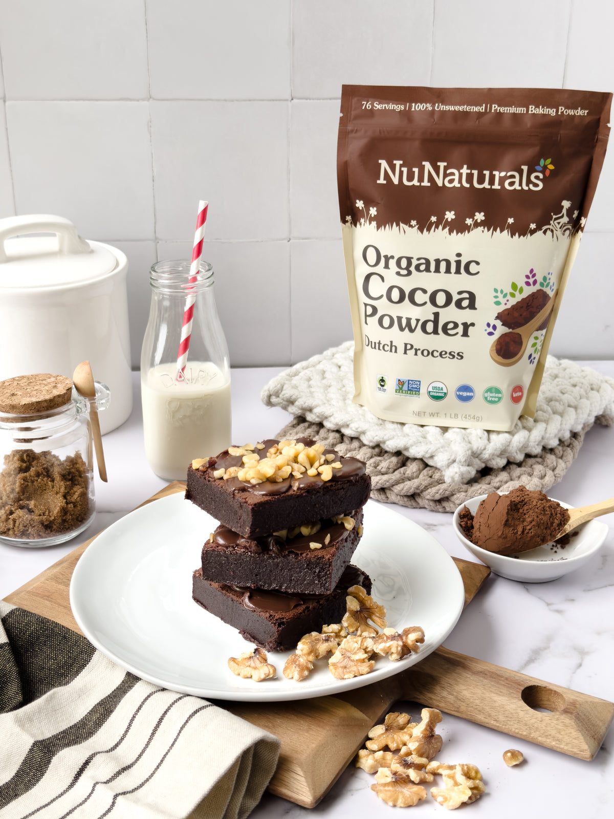 Organic Dutch Cocoa pouch behind a stack of brownies topped with walnuts, with a bottle of milk and a bowl of cocoa powder