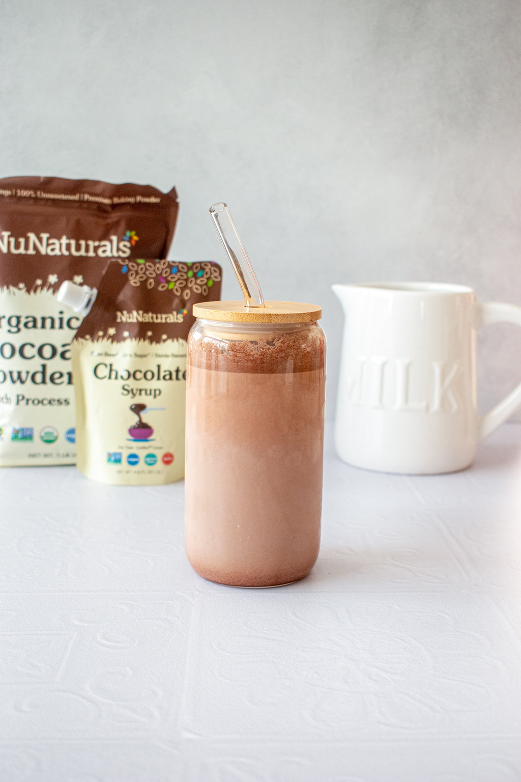 Organic Dutch Cocoa Powder pouch next to a Chocolate Syrup pouch behind a glass of chocolate milk with a glass straw