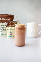 Organic Dutch Cocoa Powder pouch next to a Chocolate Syrup pouch behind a glass of chocolate milk with a glass straw