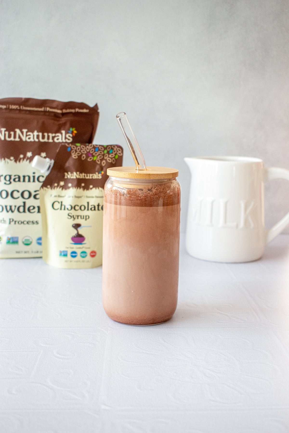 Organic Dutch Cocoa Powder pouch next to a Chocolate Syrup pouch behind a glass of chocolate milk with a glass straw