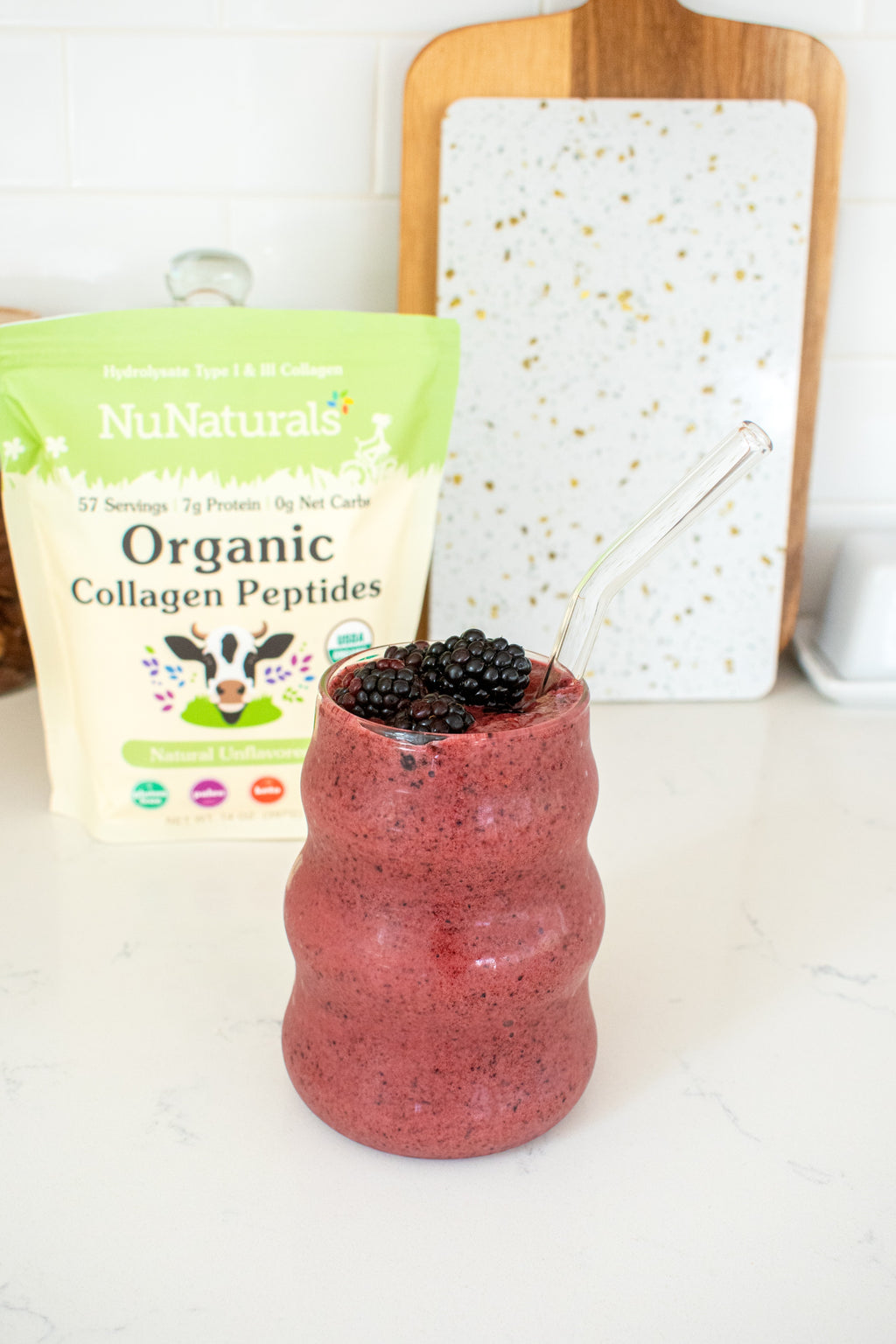 Organic Collagen Peptides pouch behind a glass of blackberry smoothie with a glass straw