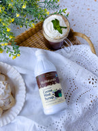 Cocoa Mint Syrup 16 oz bottle lying on white eyelet cloth beside a mug topped with whipped cream and mint