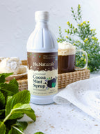 Cocoa Mint Syrup 16 oz bottle beside a glass mug of cocoa on a woven tray, with mint leaves and pastries around it