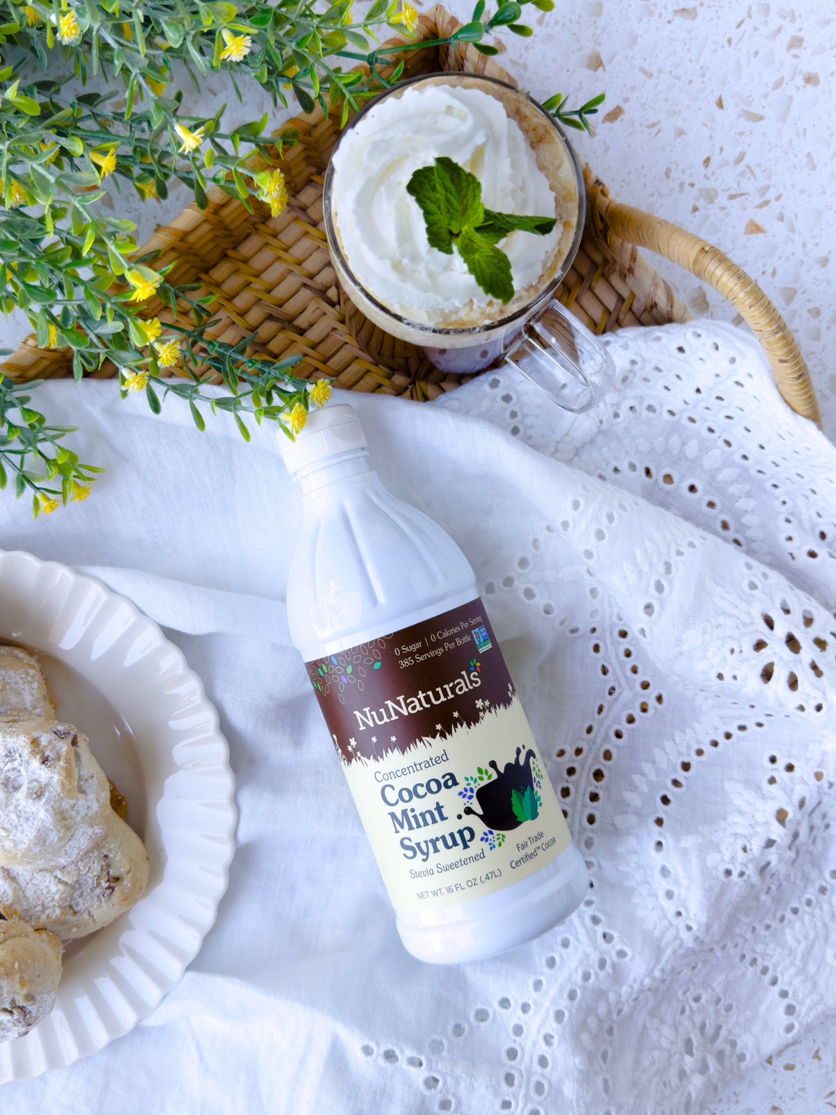 Cocoa Mint Syrup 16 oz bottle lying on white lace fabric beside a glass topped with whipped cream and a mint leaf