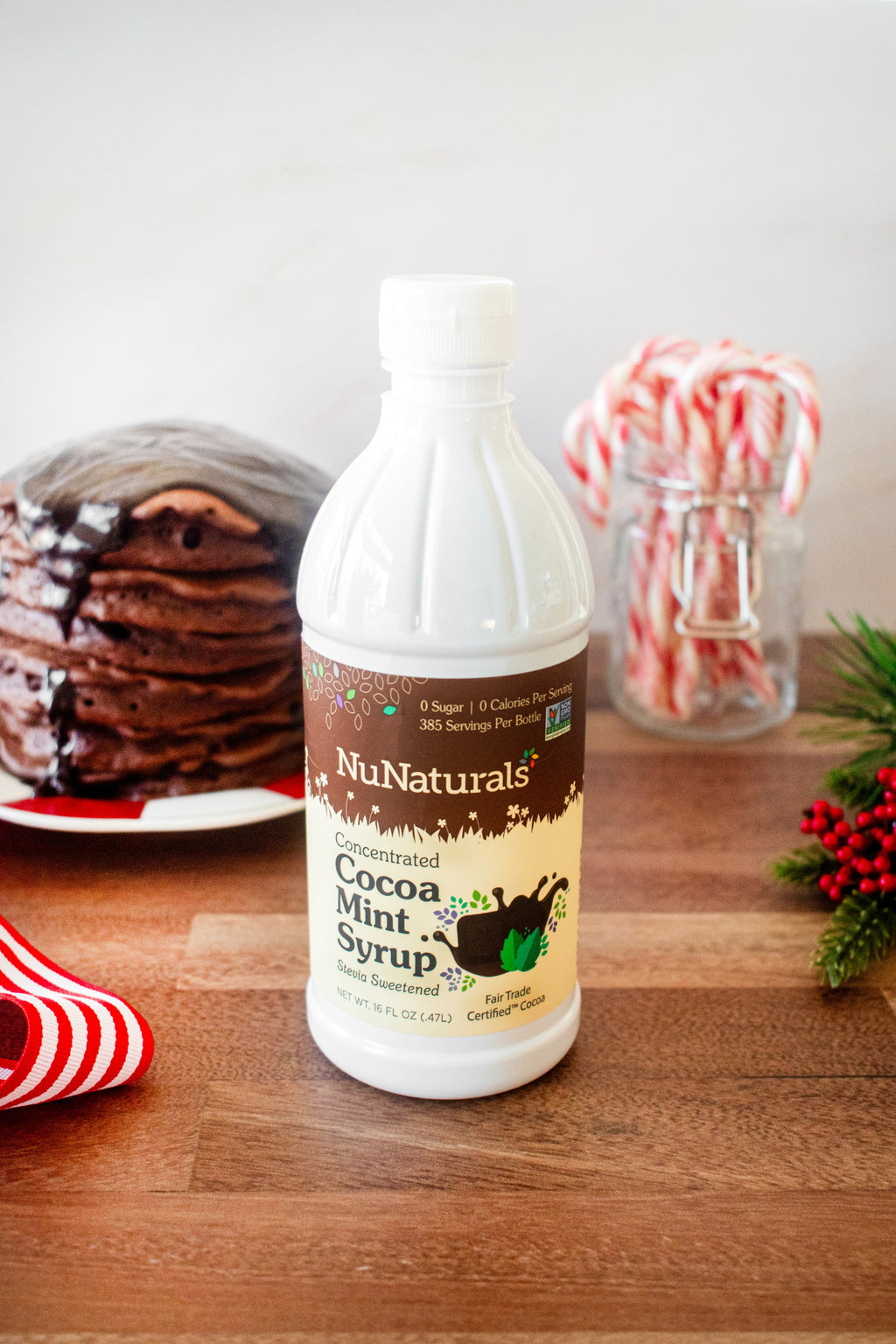 Cocoa Mint Syrup 16 oz bottle on a wooden counter with a stack of pancakes, candy canes in a jar, and holly