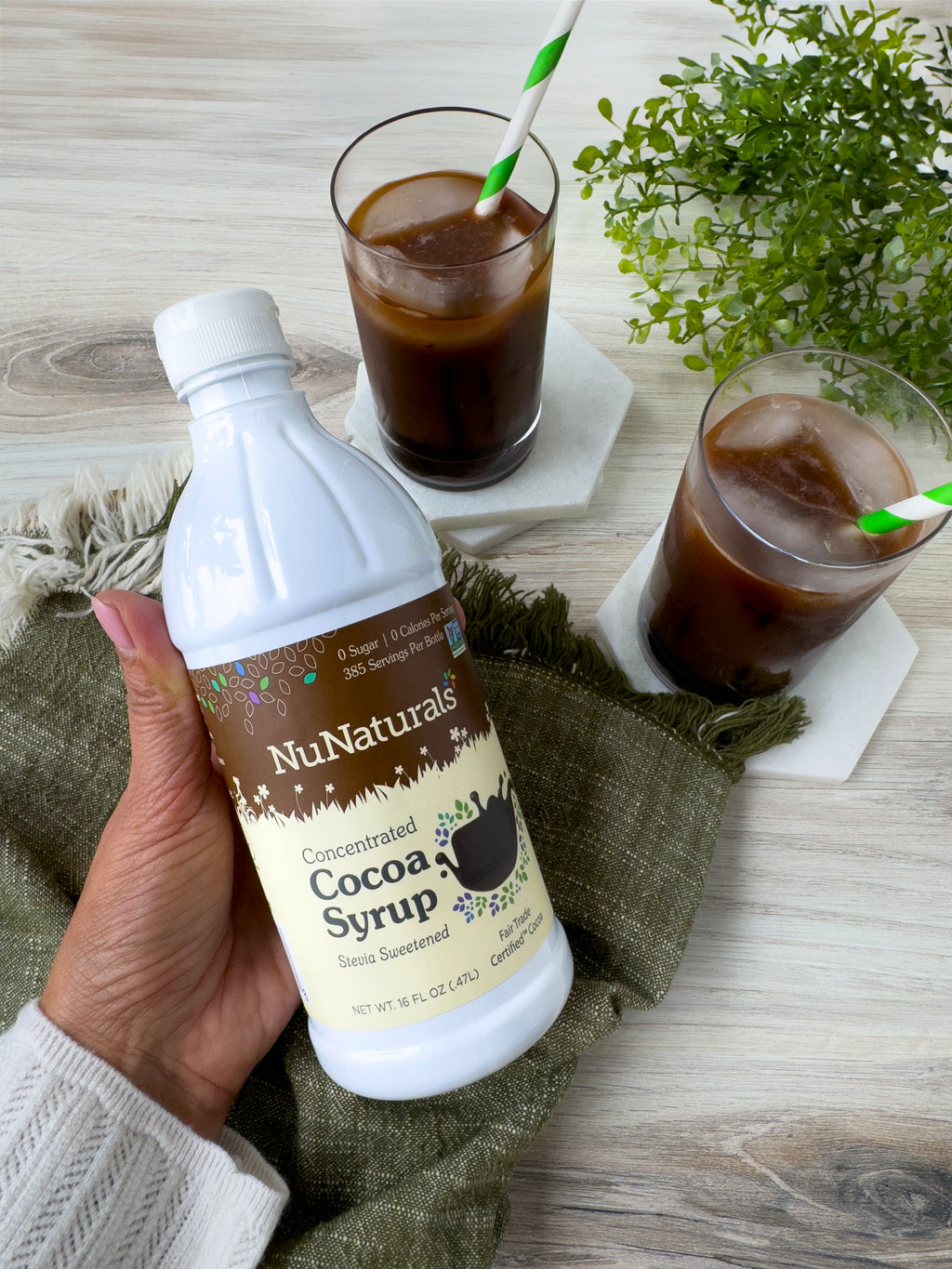 Hand holding a Cocoa Syrup 16 oz bottle over a table with two iced chocolate drinks in glasses with green straws