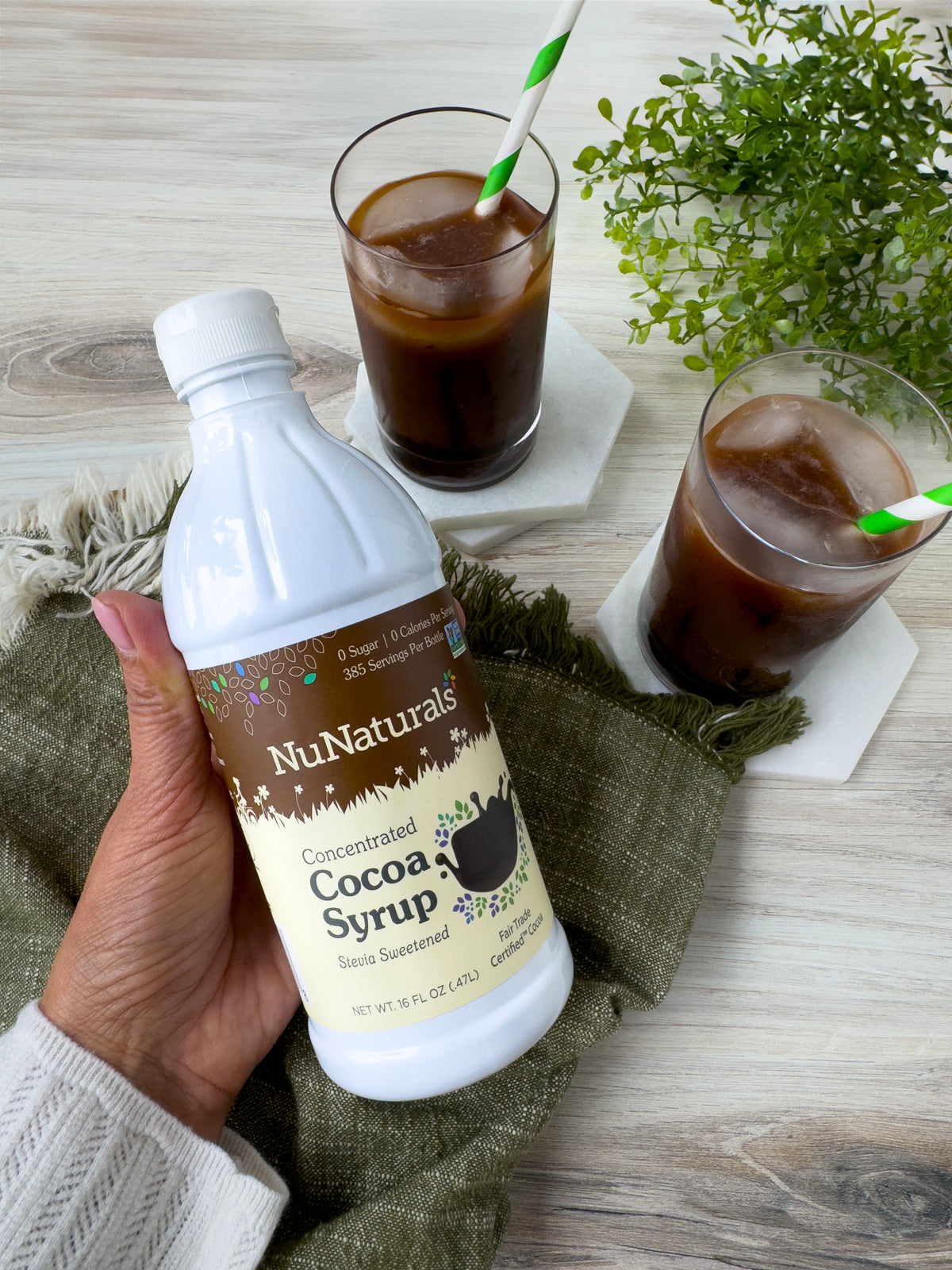 Hand holding a Cocoa Syrup 16 oz bottle over a table with two iced chocolate drinks in glasses with green straws