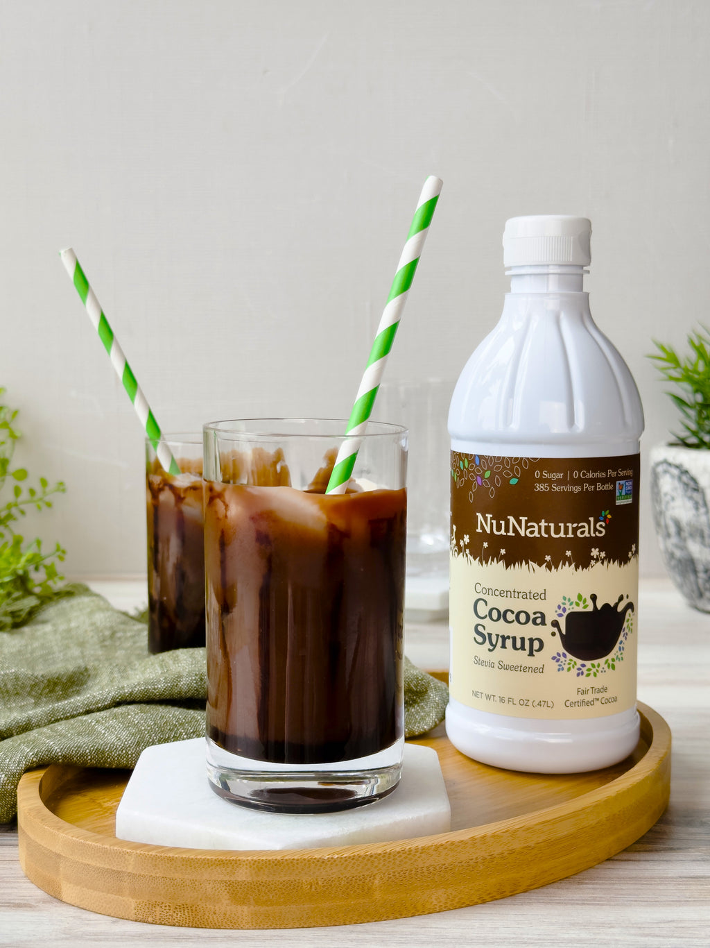 Cocoa Syrup 16 oz bottle beside two glasses of iced chocolate drink with striped paper straws on a wooden tray