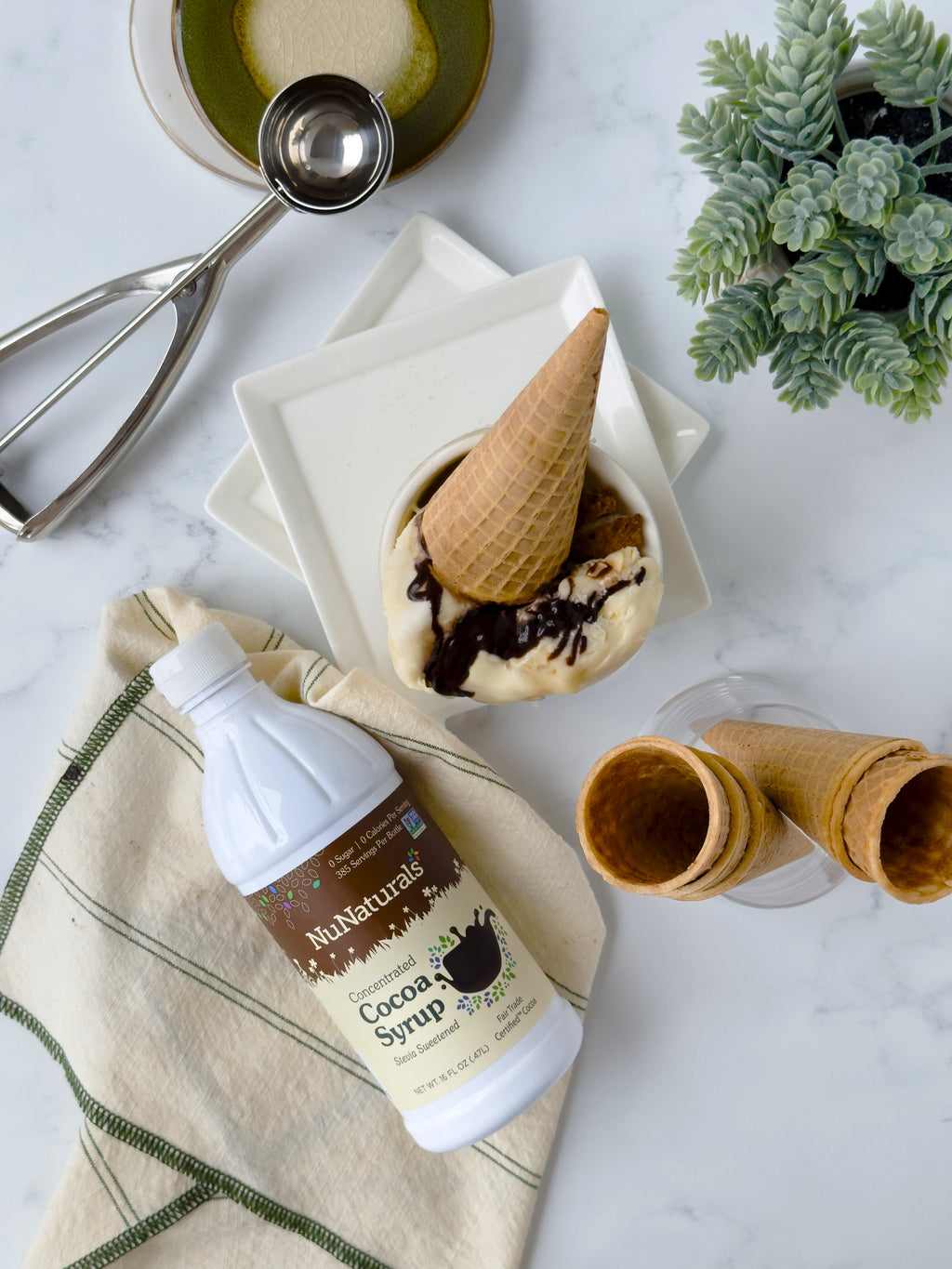 Cocoa Syrup 16 oz bottle on a folded towel beside an ice cream cone in a dish, cones and an ice cream scoop nearby
