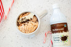 Cocoa Syrup 16 oz bottle at the edge of the frame beside a mug of dessert topped with chocolate sprinkles and a candy cane