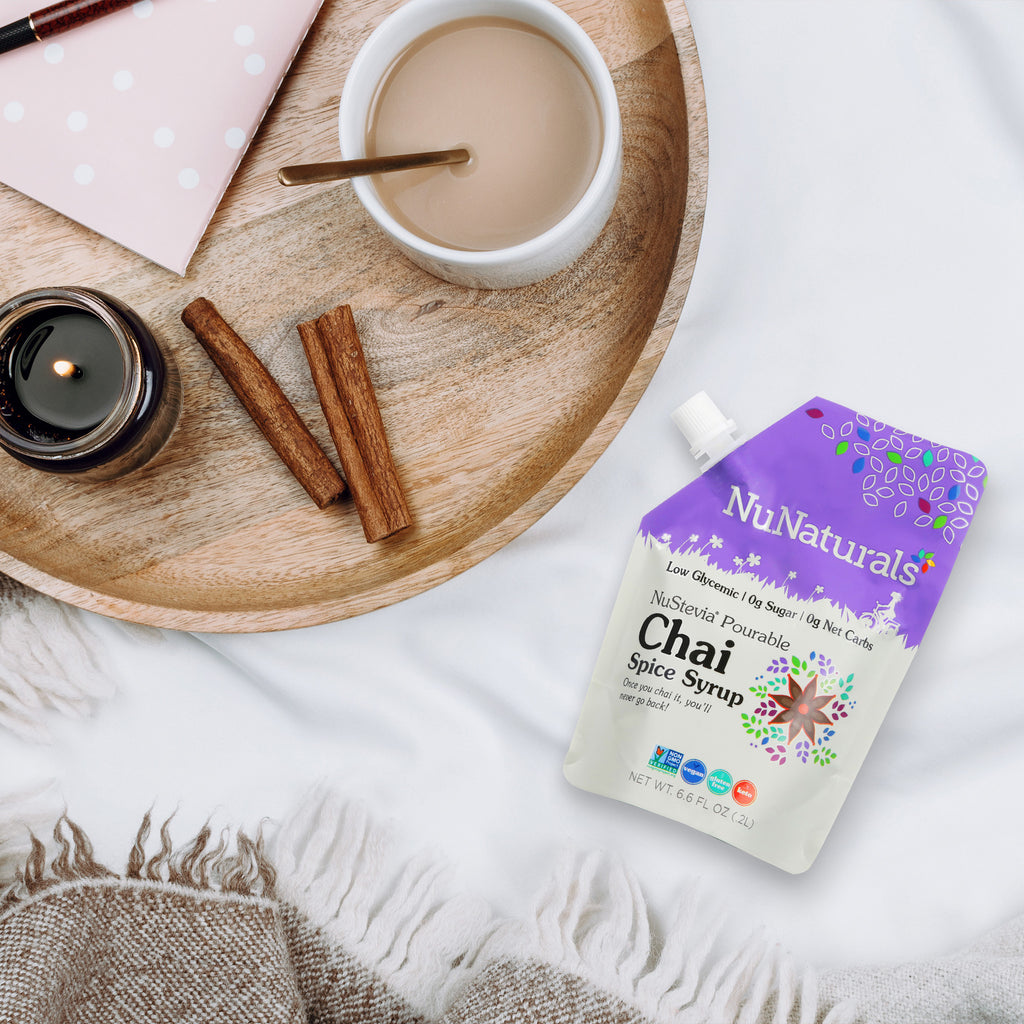 Chai Spice Syrup 6.6 oz pouch on a white blanket beside a wooden tray with a cup of tea, cinnamon sticks and a candle