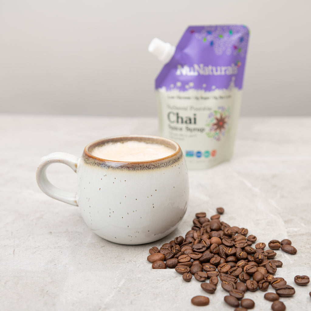 Chai Spice Syrup 6.6 oz pouch behind a ceramic mug of coffee drink, with coffee beans on a stone counter