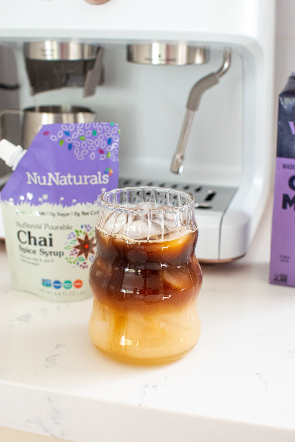 Chai Spice Syrup 6.6 oz pouch beside an iced coffee drink in a ridged glass, with an espresso machine behind