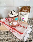 Chocolate Syrup 6.6 oz pouch beside a heart-decorated mug of whipped cream drink on a checked towel