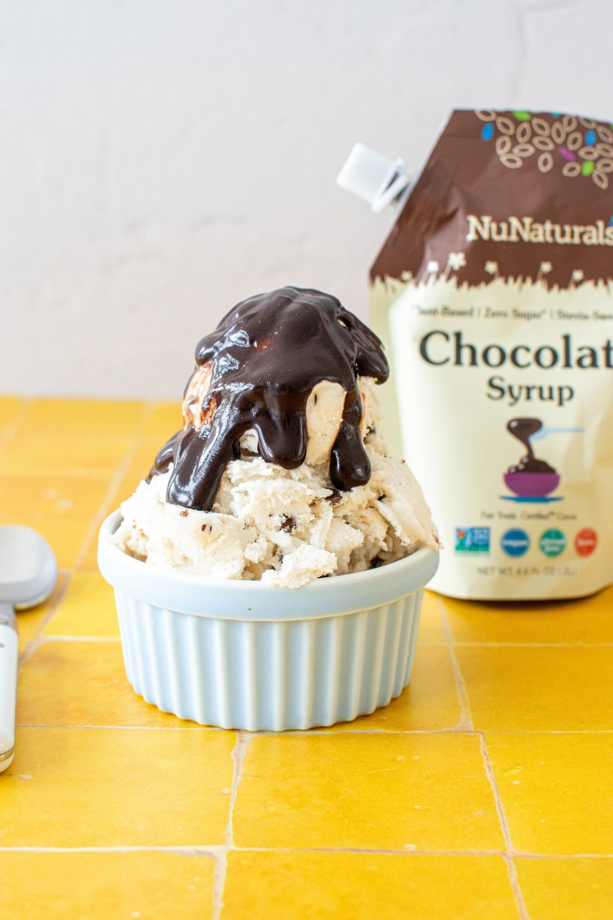 Chocolate Syrup 6.6 oz pouch behind a bowl of ice cream topped with syrup, on a yellow tiled surface