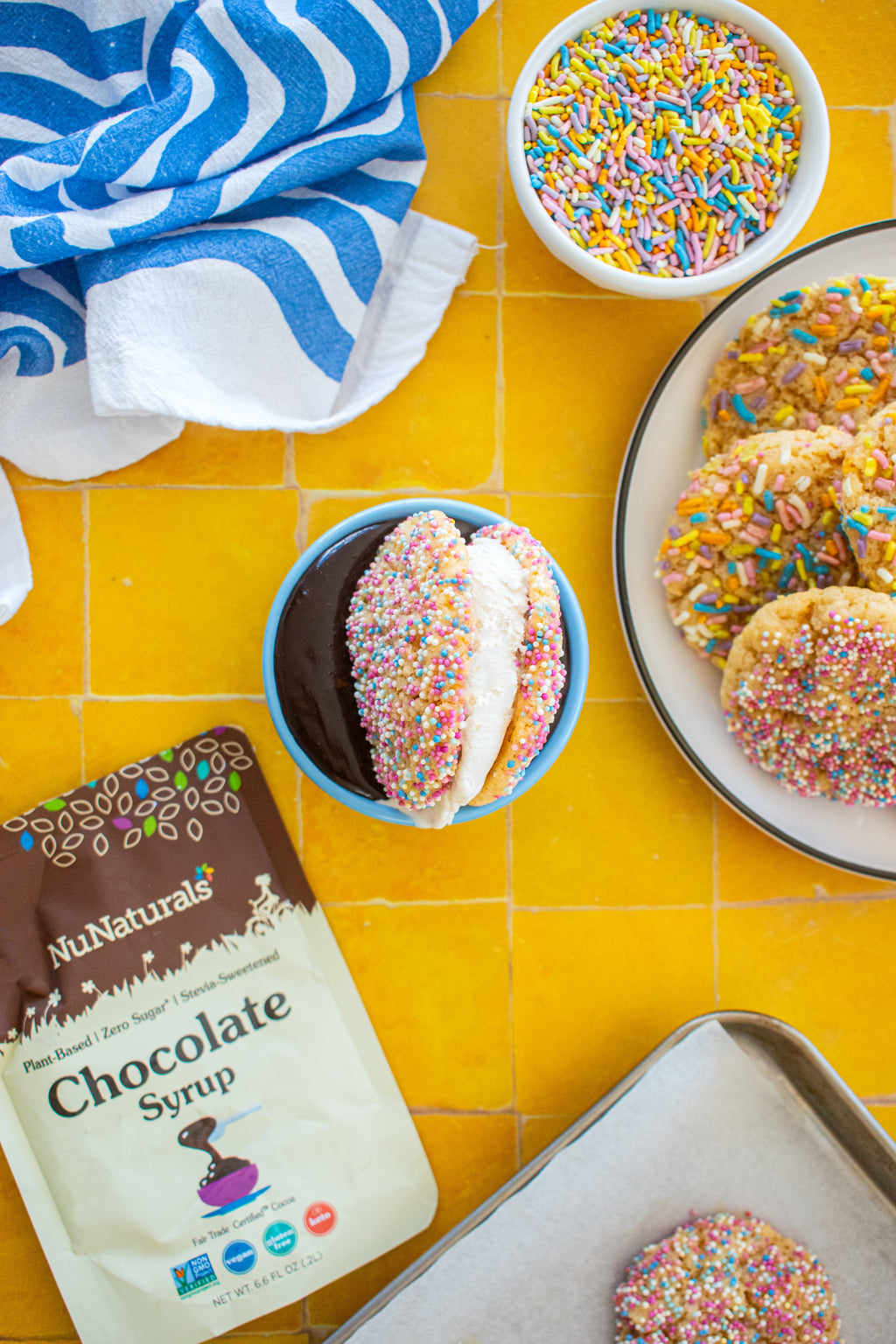 Chocolate Syrup 6.6 oz pouch at the corner of a yellow tile counter beside a bowl of dip and sprinkle-coated cookies