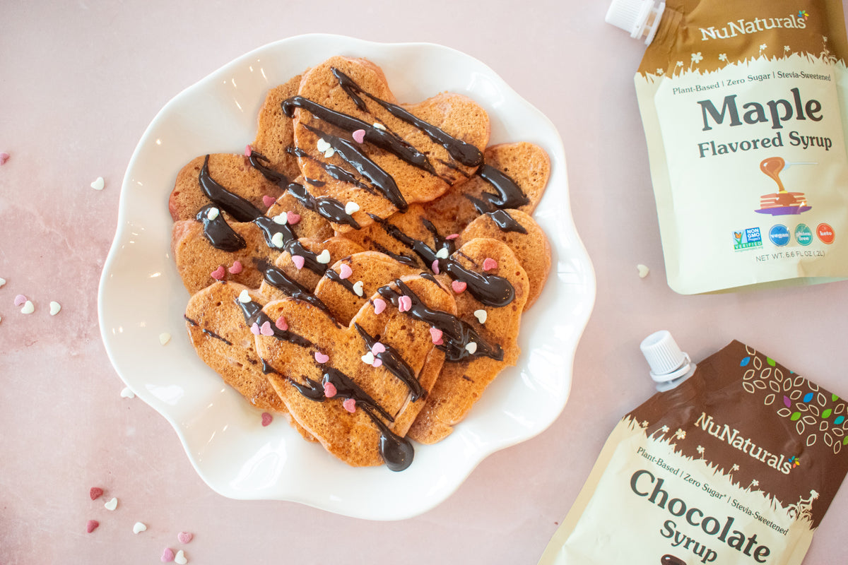 Heart-shaped pancakes drizzled with chocolate syrup on a white plate, with Chocolate Syrup 6.6 oz pouches at the edges