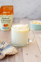 Salted Caramel Flavored Syrup 6.6 oz pouch beside a glass mug of drink topped with cream, with a bowl of nuts behind