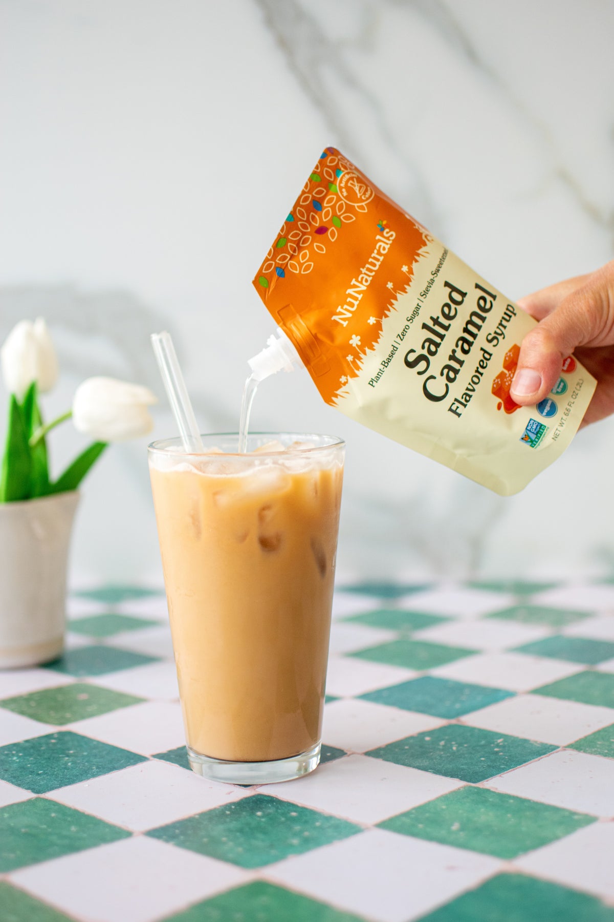 Salted Caramel Flavored Syrup 6.6 oz pouch held by a hand, pouring into an iced coffee drink with a straw