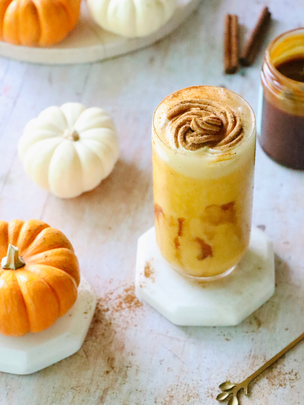 Cup of orange pumpkin drink topped with cinnamon cream, beside small pumpkins and a jar