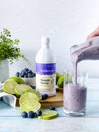 Simple Syrup 16 oz bottle on a cutting board with limes and blueberries, and a hand pouring a purple smoothie into a glass