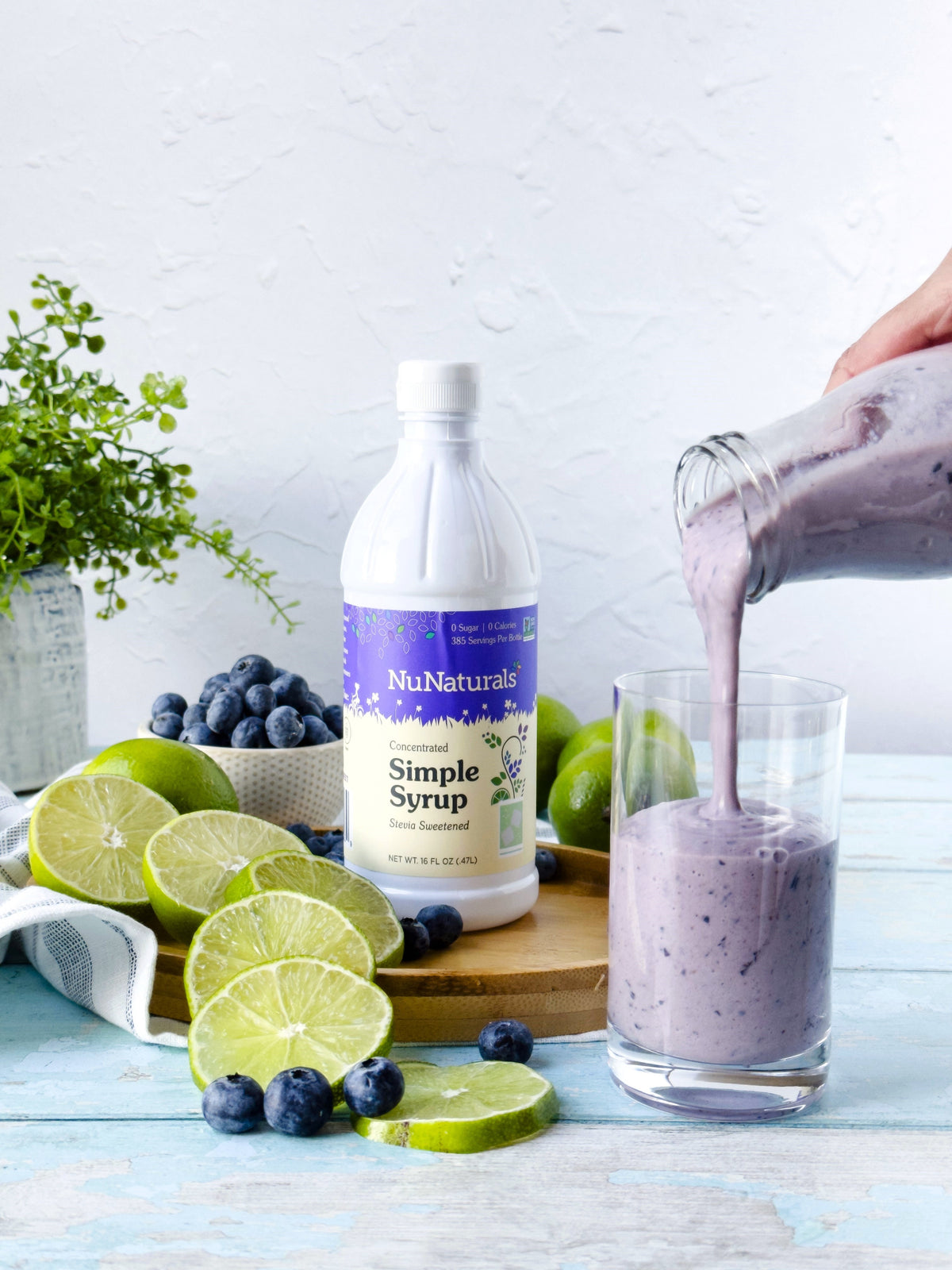 Simple Syrup 16 oz bottle on a cutting board with limes and blueberries, and a hand pouring a purple smoothie into a glass