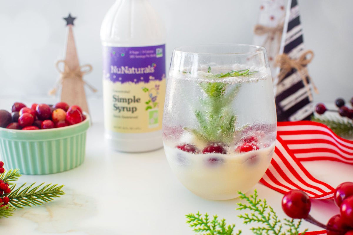 Simple Syrup 16 oz bottle behind a stemless glass of cranberries and a sprig of evergreen, with holiday decor around it