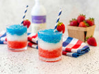 Simple Syrup 16 oz bottle out of focus behind two layered red, white, and blue frozen drinks with strawberries