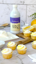 Vanilla Syrup 16 oz bottle beside lemon-topped cupcakes with yellow and white frosting, lemons and a whisk behind