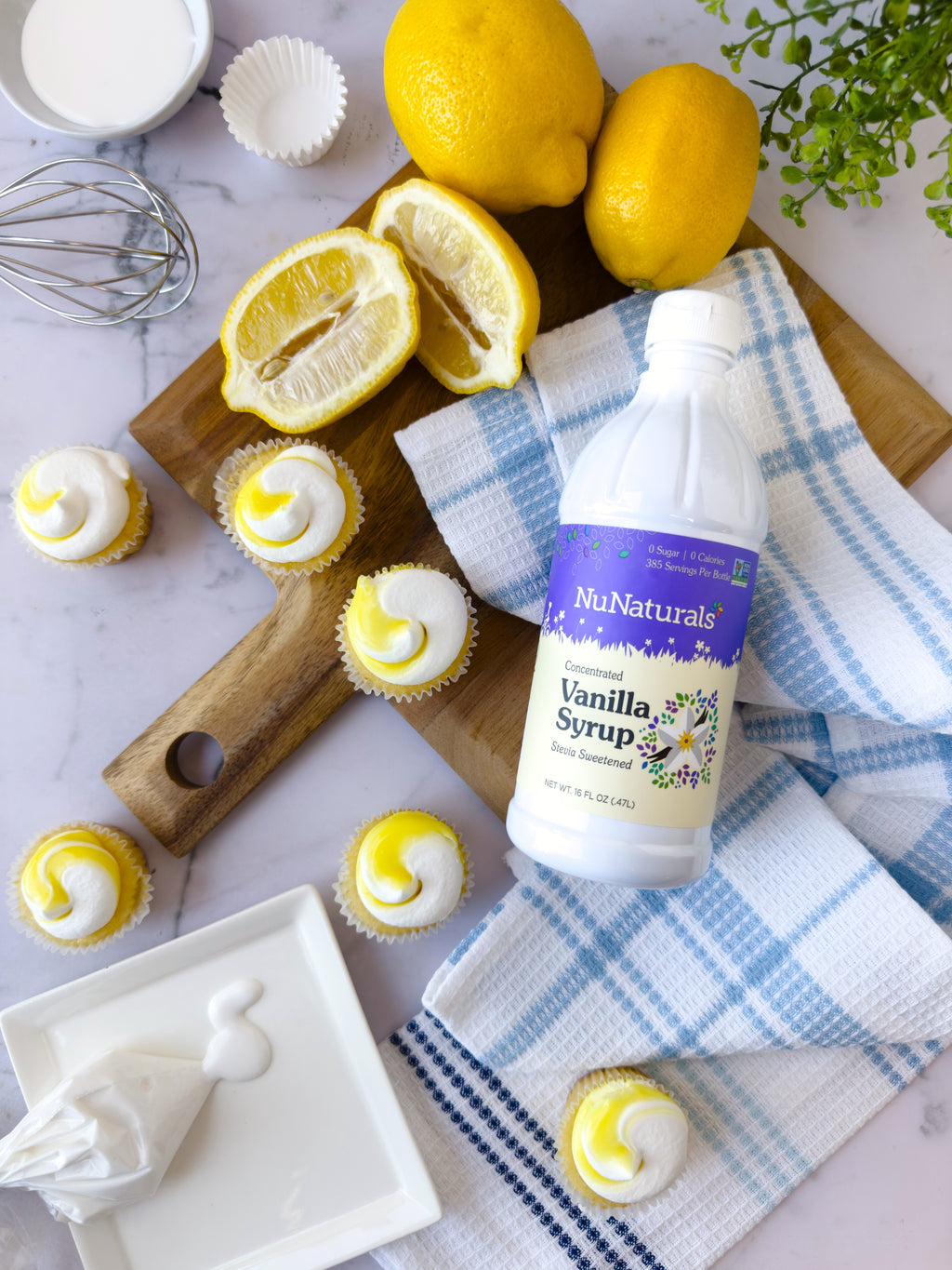 Vanilla Syrup 16 oz bottle lying on a blue checked towel beside lemons and lemon cupcakes with white frosting