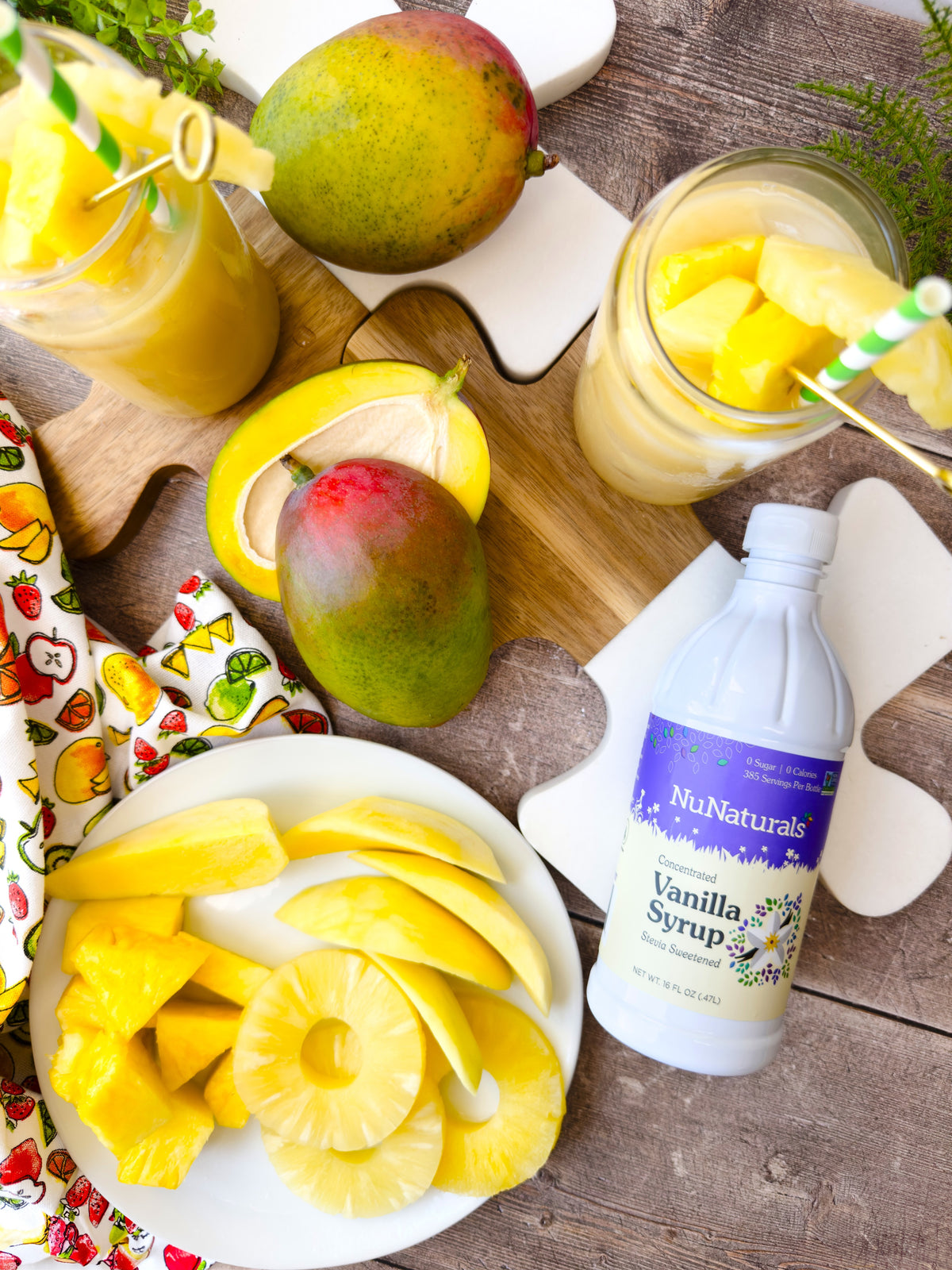 Vanilla Syrup 16 oz bottle on a wooden board beside sliced mango, pineapple rings, and glasses of fruit drink