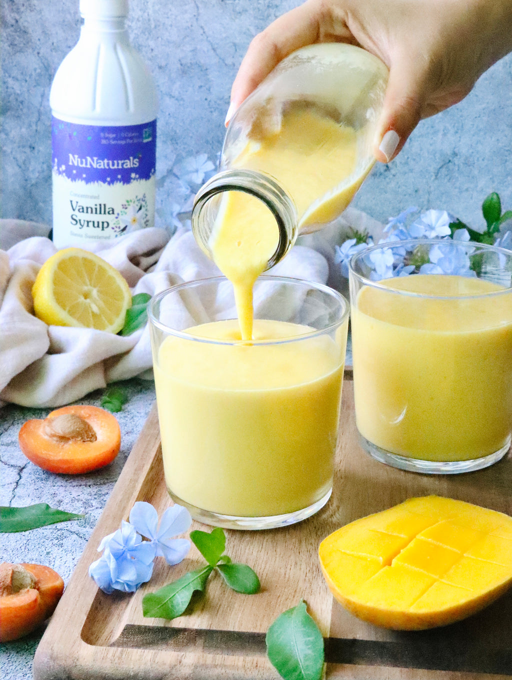 Vanilla Syrup 16 oz bottle in the corner behind a hand pouring a yellow smoothie into glasses, with lemon and mango