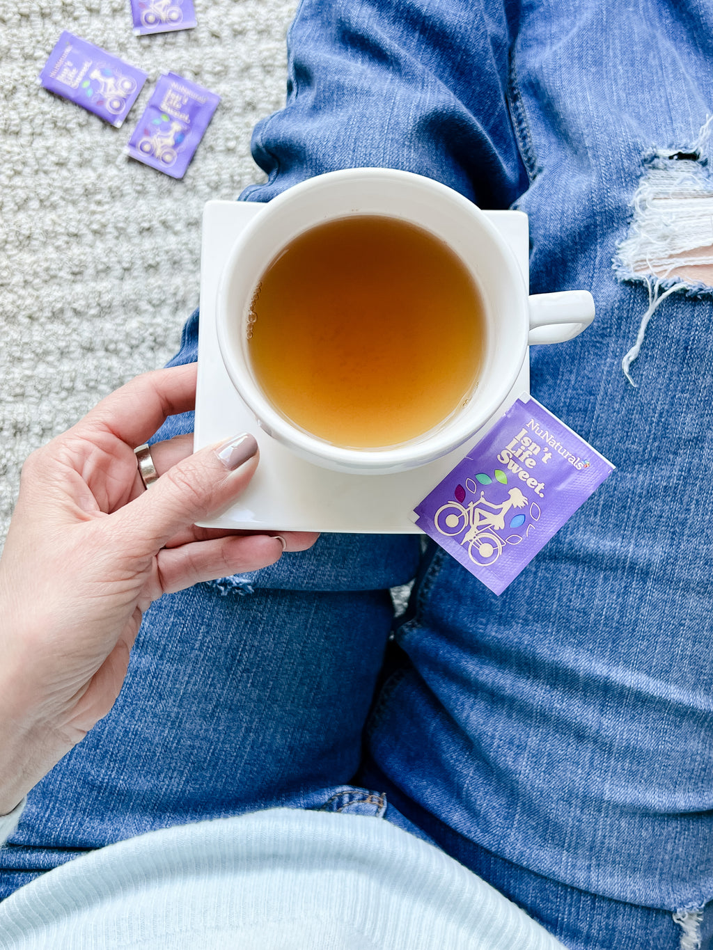 White Stevia Powder Blend Packets tea bag style packet resting on a saucer beside a cup of tea, held over jeans