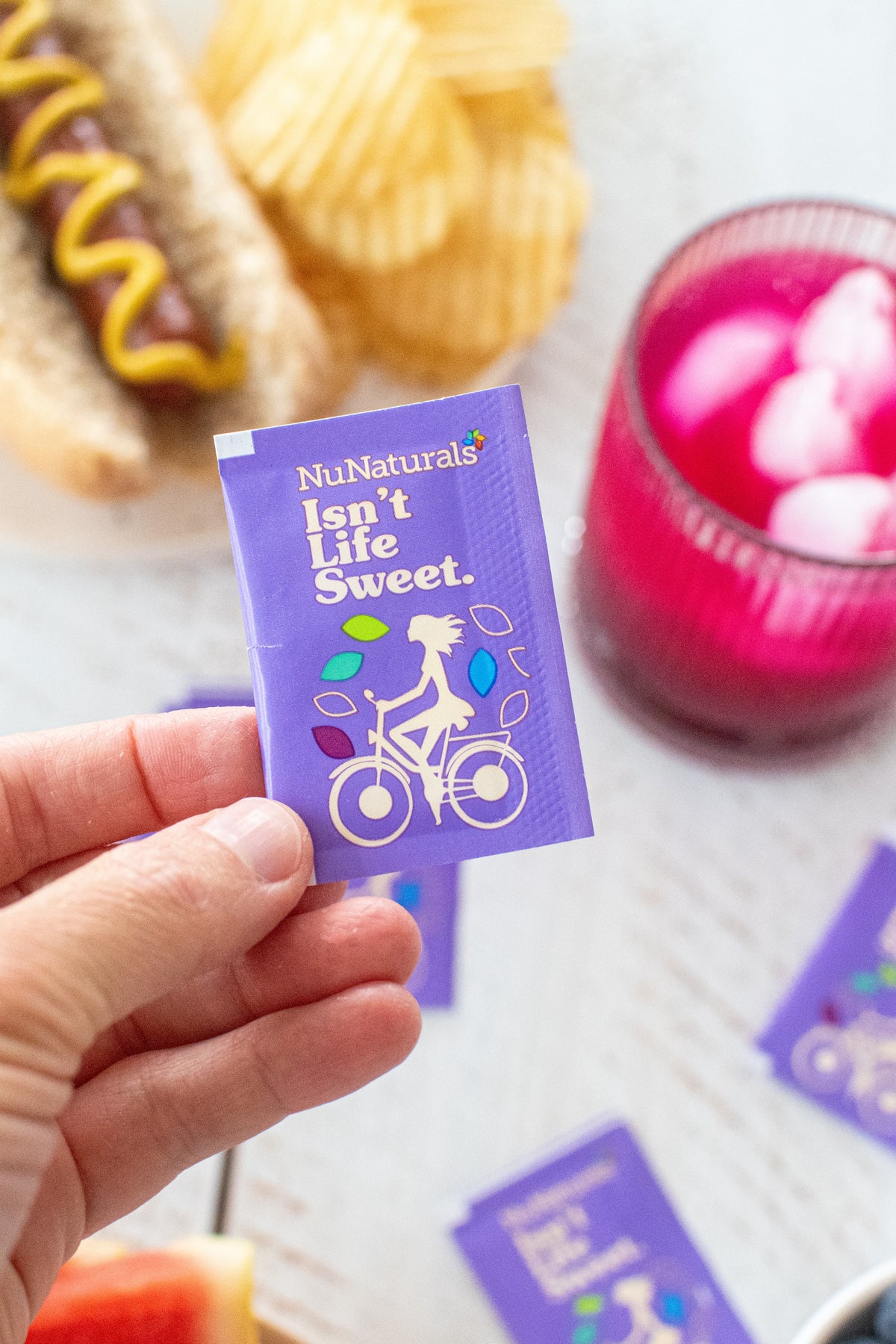 White Stevia Powder Blend Packets, one purple packet held in a hand, with a pink drink and hot dog in the background